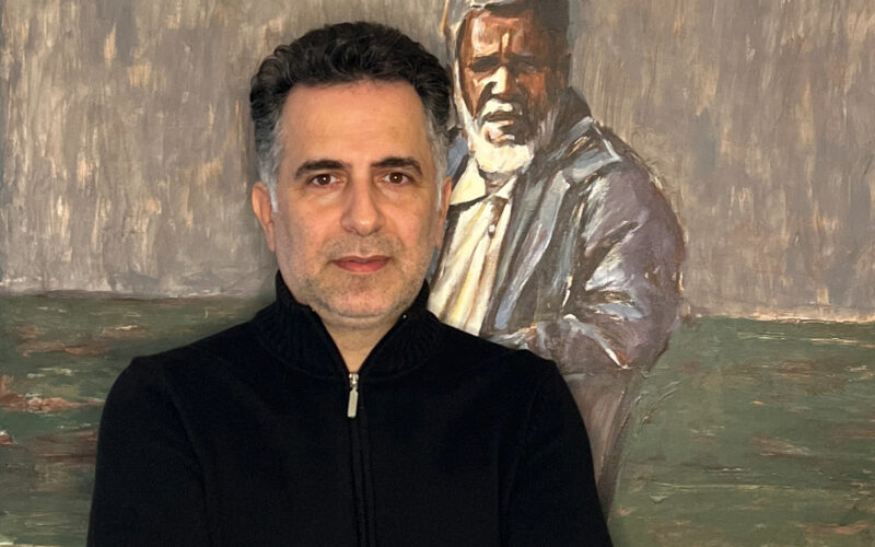 Das Foto zeigt Reza Ghanei vor einem seiner gemalten Bilder