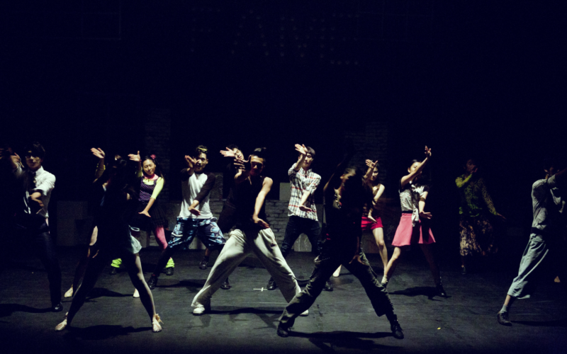 Das Bild zeigt ein großes Tanzensemble aus jungen Leuten auf einer Bühne