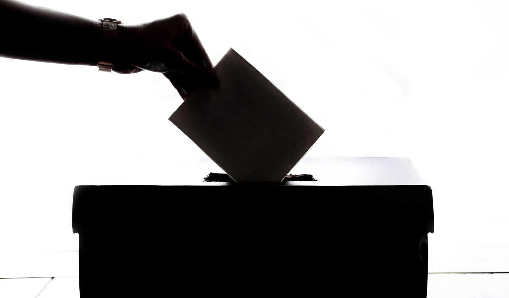 Das Bild zeigt eine Hand wie sie einen Wahlzettel in eine Wahlurne steckt. Die Hand und die Box liegen im Schatten