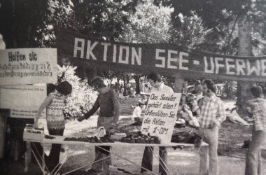 Schwarzweißfotografie aus den 1970er-Jahren. Im Vordergrund ein Infostand mit mehreren jungen Menschen unter einem großen Transparent mit der Aufschrift „AKTION SEE-UFERWEG“. Auf einem Schild steht: „Das Seeufer gehört allen! Unterstützen Sie die Aktion mit 1,- DM“. Im Hintergrund sitzen Menschen auf einer Wiese im Schatten von Bäumen. Die Szene zeigt eine Spendensammelaktion für den öffentlichen Zugang zum Konstanzer Seeufer, vermutlich im Rahmen der Proteste von 1975.