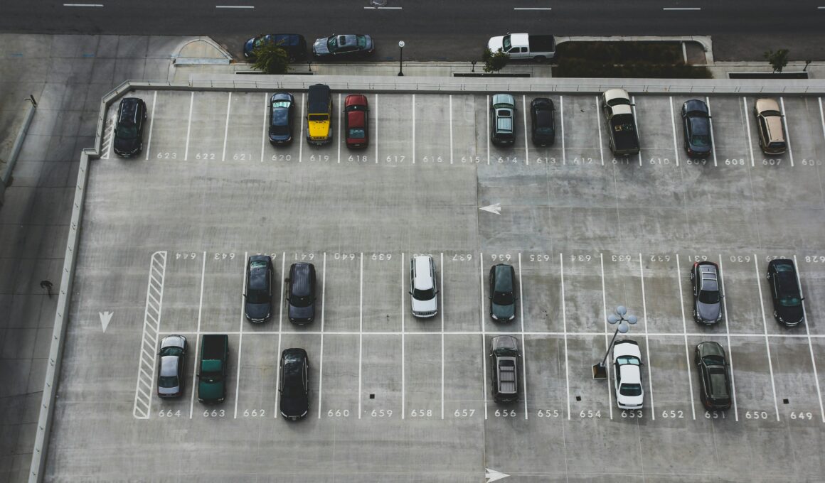 Luftaufnahme eines halb gefüllten Parkdecks mit markierten Stellplätzen und parkenden Autos. Im oberen Bildbereich verläuft eine mehrspurige Straße.