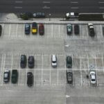 Luftaufnahme eines halb gefüllten Parkdecks mit markierten Stellplätzen und parkenden Autos. Im oberen Bildbereich verläuft eine mehrspurige Straße.