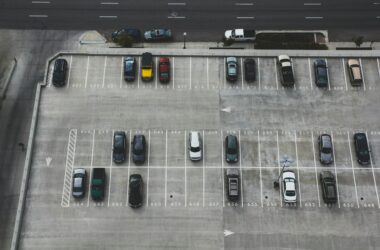 Luftaufnahme eines halb gefüllten Parkdecks mit markierten Stellplätzen und parkenden Autos. Im oberen Bildbereich verläuft eine mehrspurige Straße.