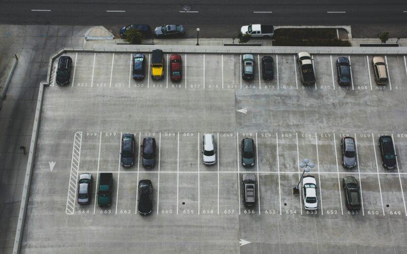 Luftaufnahme eines halb gefüllten Parkdecks mit markierten Stellplätzen und parkenden Autos. Im oberen Bildbereich verläuft eine mehrspurige Straße.