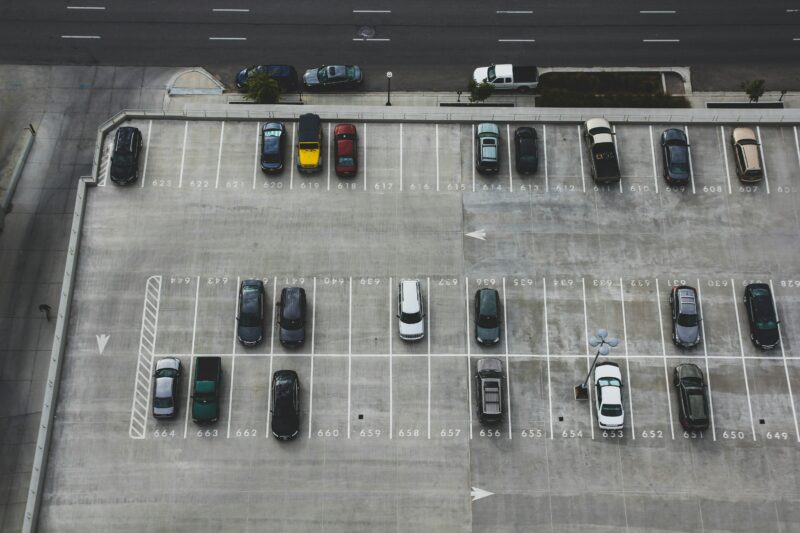 Luftaufnahme eines halb gefüllten Parkdecks mit markierten Stellplätzen und parkenden Autos. Im oberen Bildbereich verläuft eine mehrspurige Straße.
