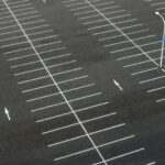 Luftaufnahme eines leeren Parkplatzes mit klar markierten Stellflächen und Richtungsmarkierungen. In der Mitte steht eine einzelne Straßenlaterne.