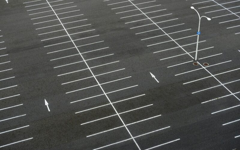 Luftaufnahme eines leeren Parkplatzes mit klar markierten Stellflächen und Richtungsmarkierungen. In der Mitte steht eine einzelne Straßenlaterne.