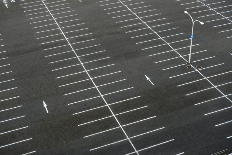 Luftaufnahme eines leeren Parkplatzes mit klar markierten Stellflächen und Richtungsmarkierungen. In der Mitte steht eine einzelne Straßenlaterne.